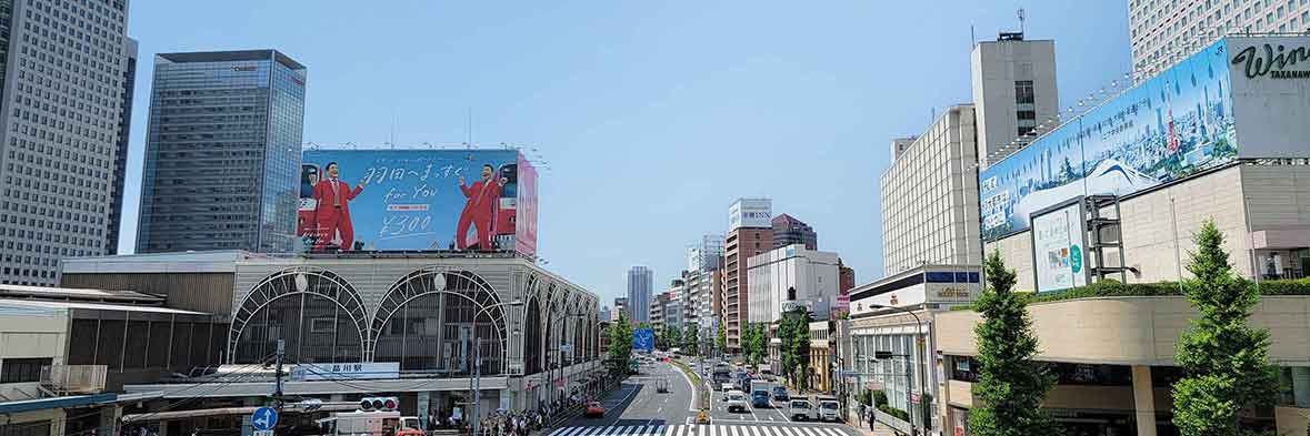 青空のもと、高層ビルが立ち並ぶ品川駅前の広々とした街並み。多くの車が行き交い、活気あふれる日常風景。